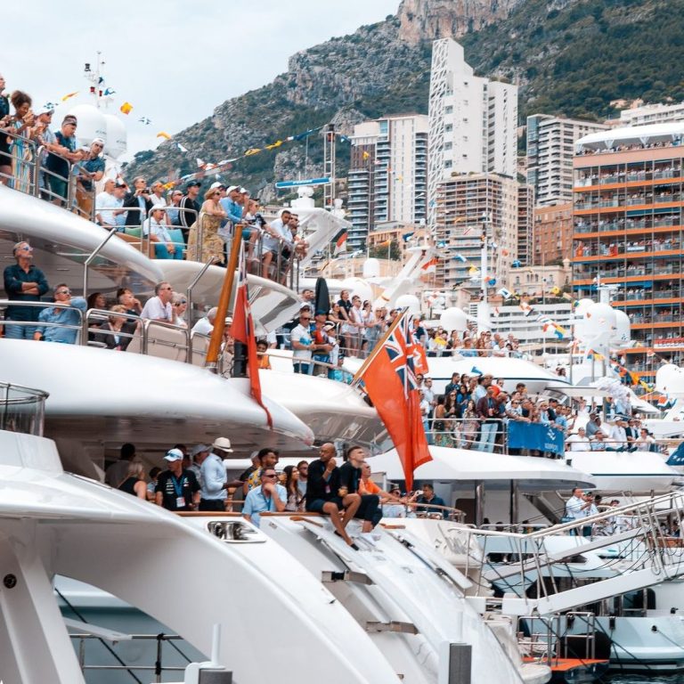 Monaco's bustling yacht harbor with crowds and mountains in the background.