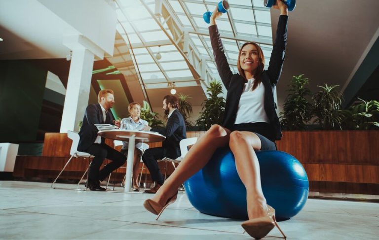 Woman sitting cross-legged on a gym ball, lifting dumbbells, in a meeting room.