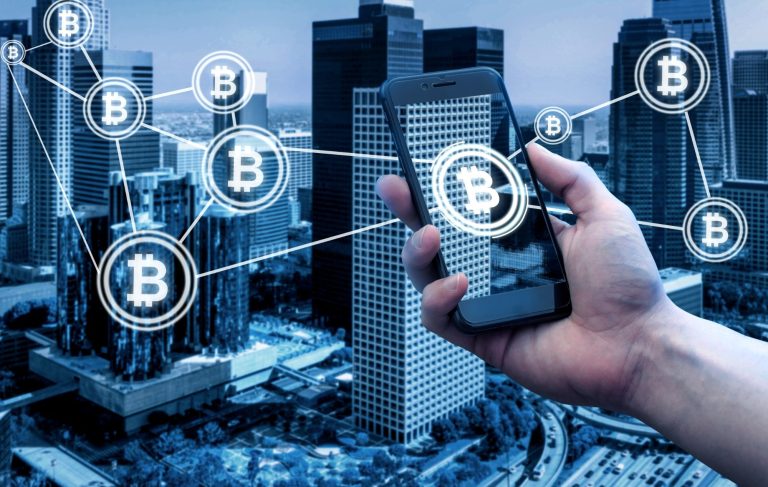 Mainly a smartphone displaying Bitcoin symbols with a city skyline in the background.