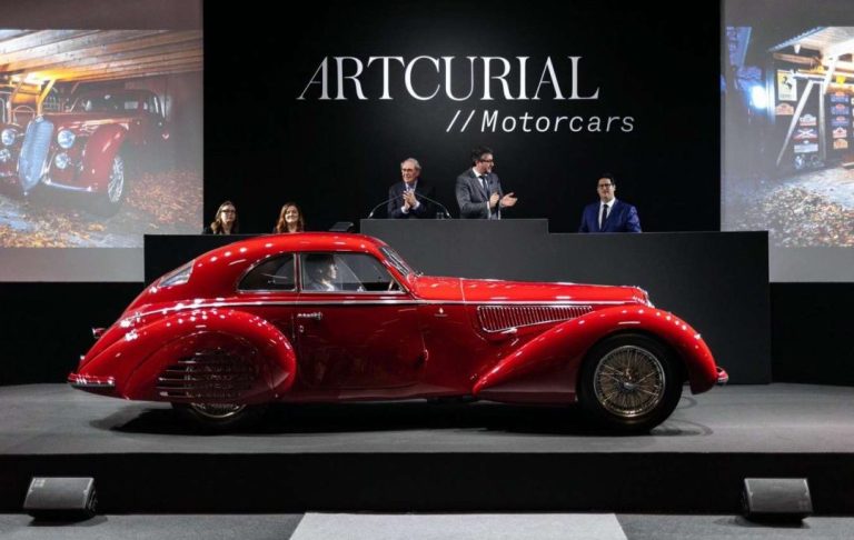 Red vintage car at auction, with podium and bidders.
