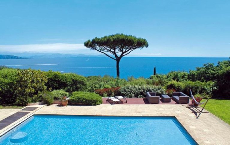 View of a garden with pool, trees and ocean in the background under a blue sky.