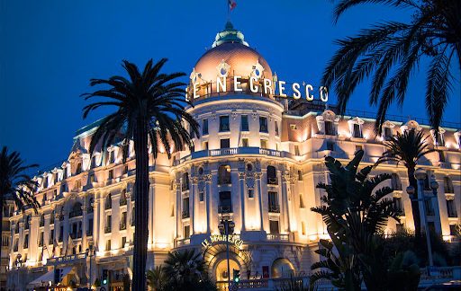Illuminated Negresco Hotel in Nice, with palm tree in foreground and night sky.