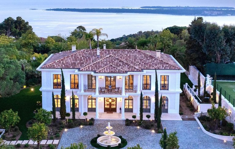 Large house with white facade, tiled roof, pool and gardens, water views.