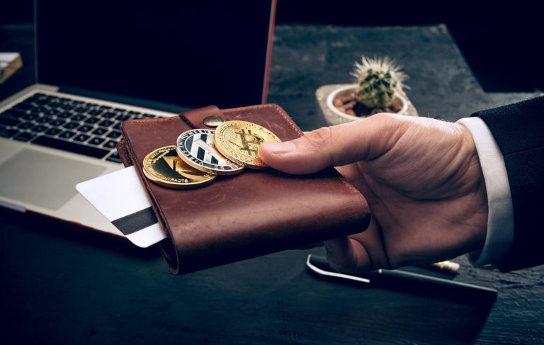 Hand in a leather wallet holding cryptocurrency coins and a smartphone on a desk.