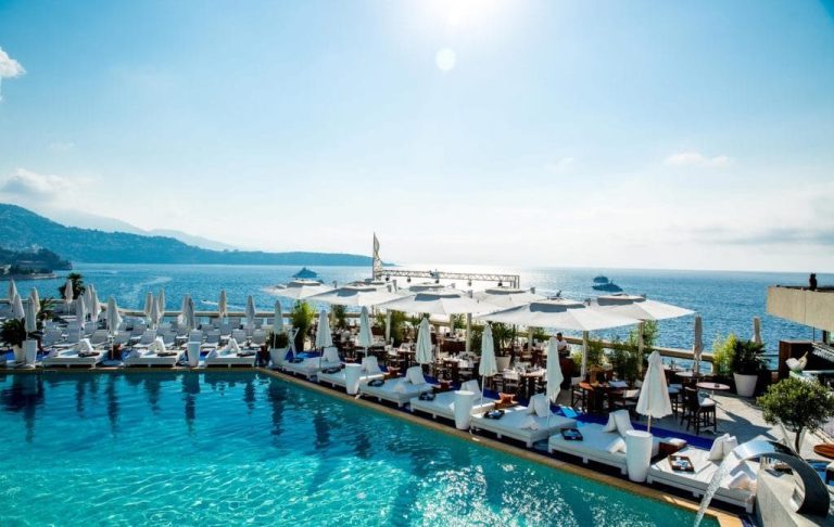 View of a pool with sun loungers and a seaside restaurant under a sunny sky.